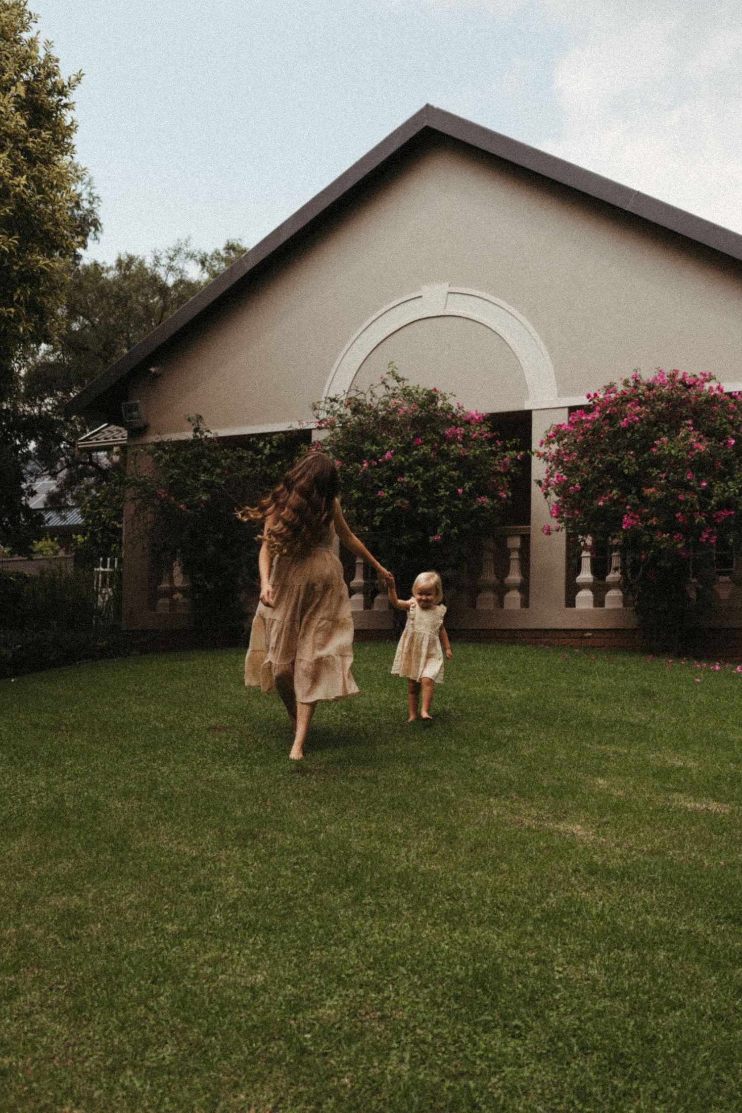 People in flowing dresses dance and twirl on a grassy lawn beside a house with blooming flowers.