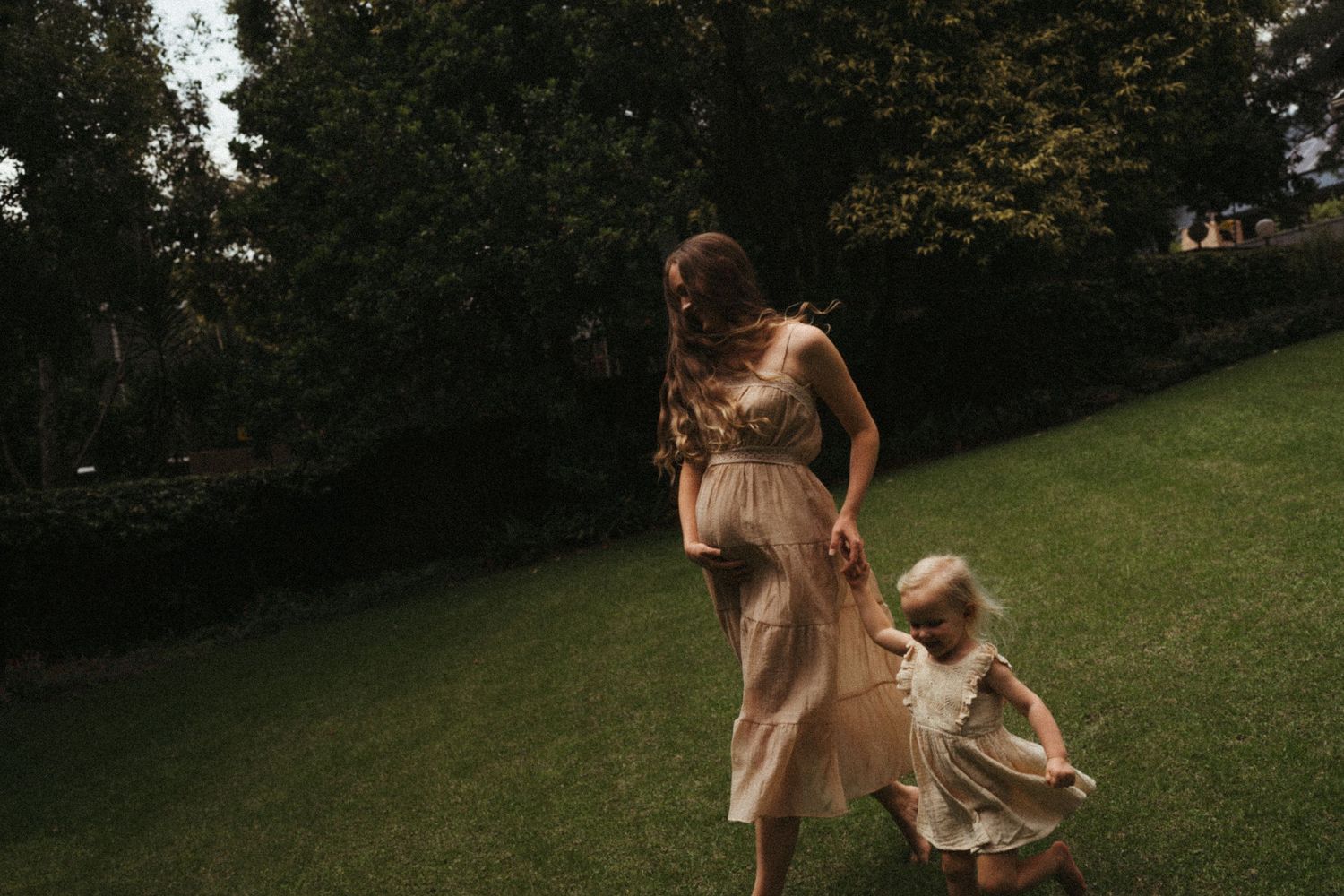 Flowing dresses catch the evening light as figures move gracefully across a lawn.