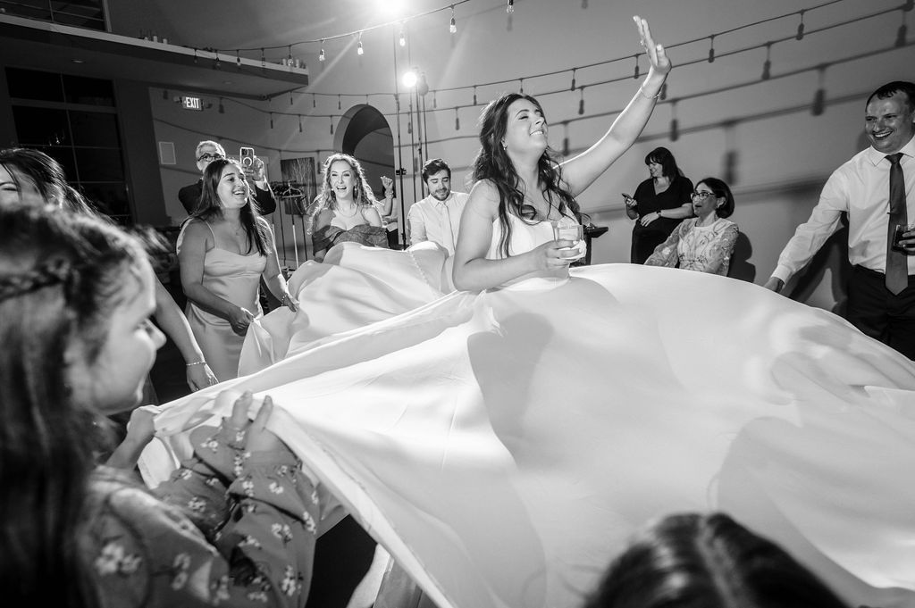 A black and white photo shows a bride's flowing veil being tossed playfully in the air during a wedding celebration.