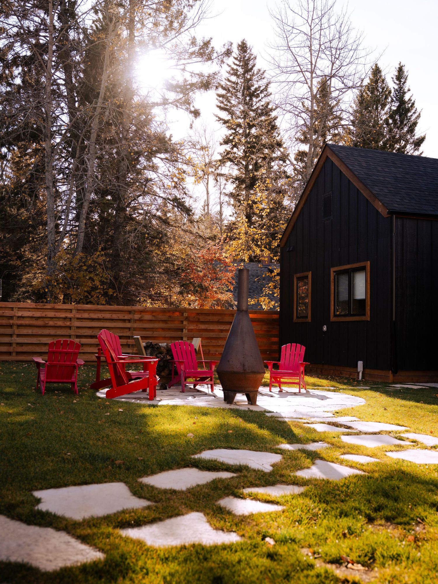 Black cabin with red Adirondack chairs and stone pathway in a sunlit forest setting.