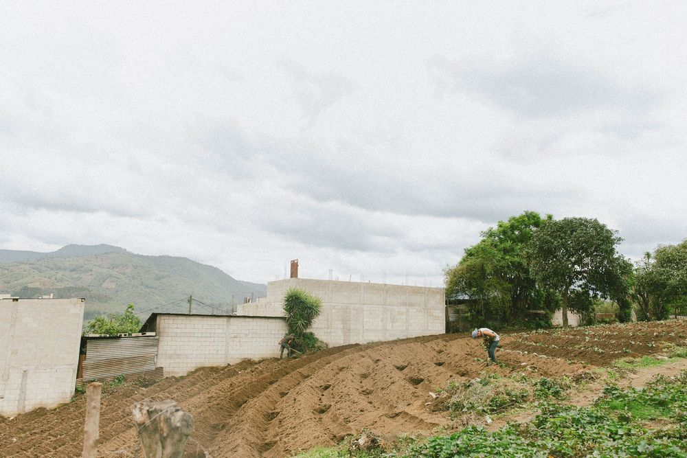 A dirt construction site on a hillside with white walls and green trees against an overcast sky.