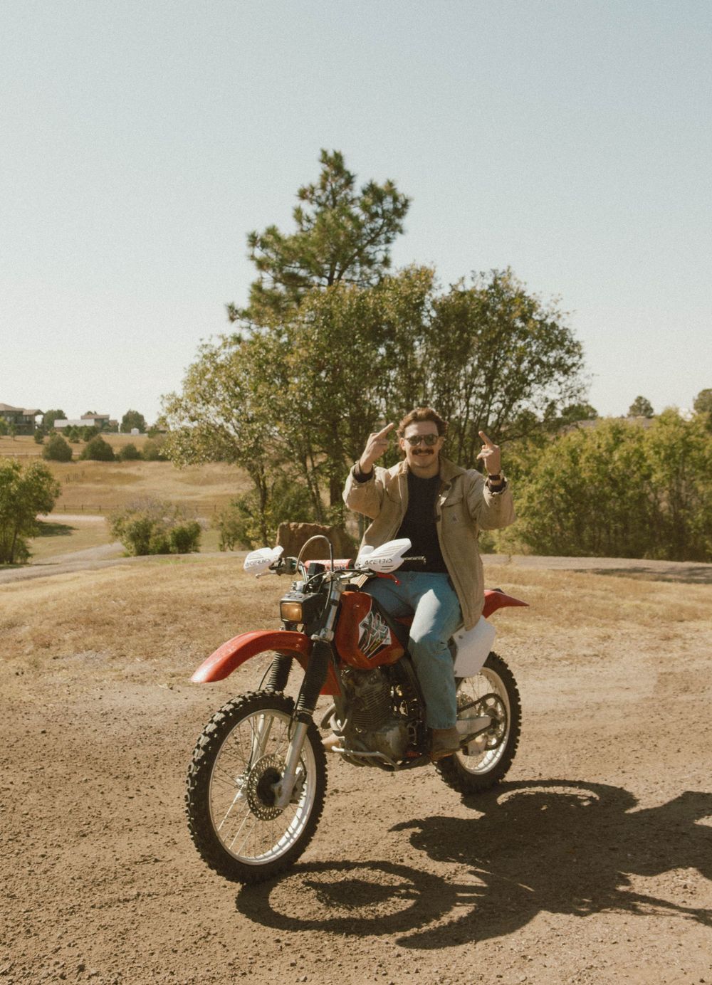 A vintage dirt bike parked on a dusty trail with trees and rolling hills in the background during golden hour.
