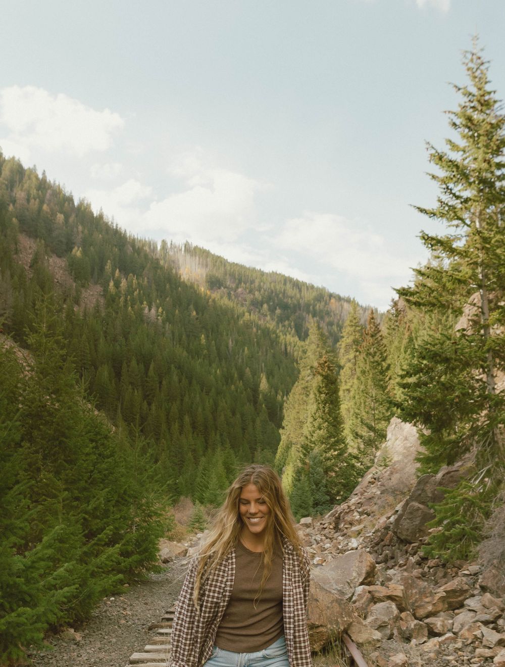 A rocky mountain trail surrounded by evergreen forest stretches into the distance under a bright sky.