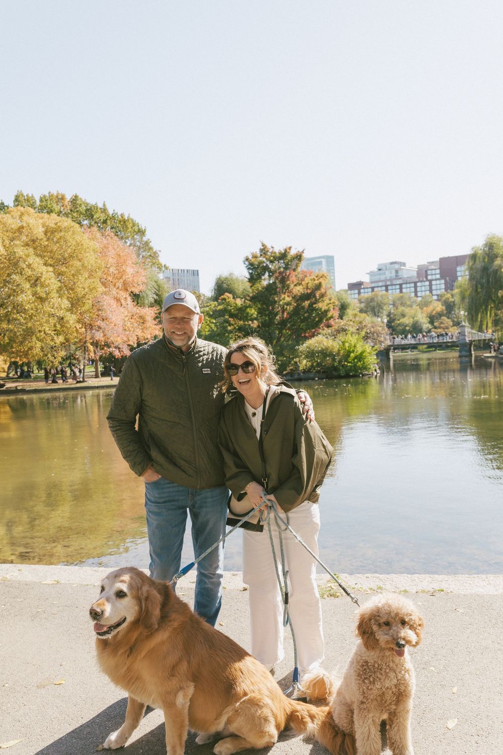 Two dogs and their owners enjoying a sunny autumn day by a pond with city buildings in the background.