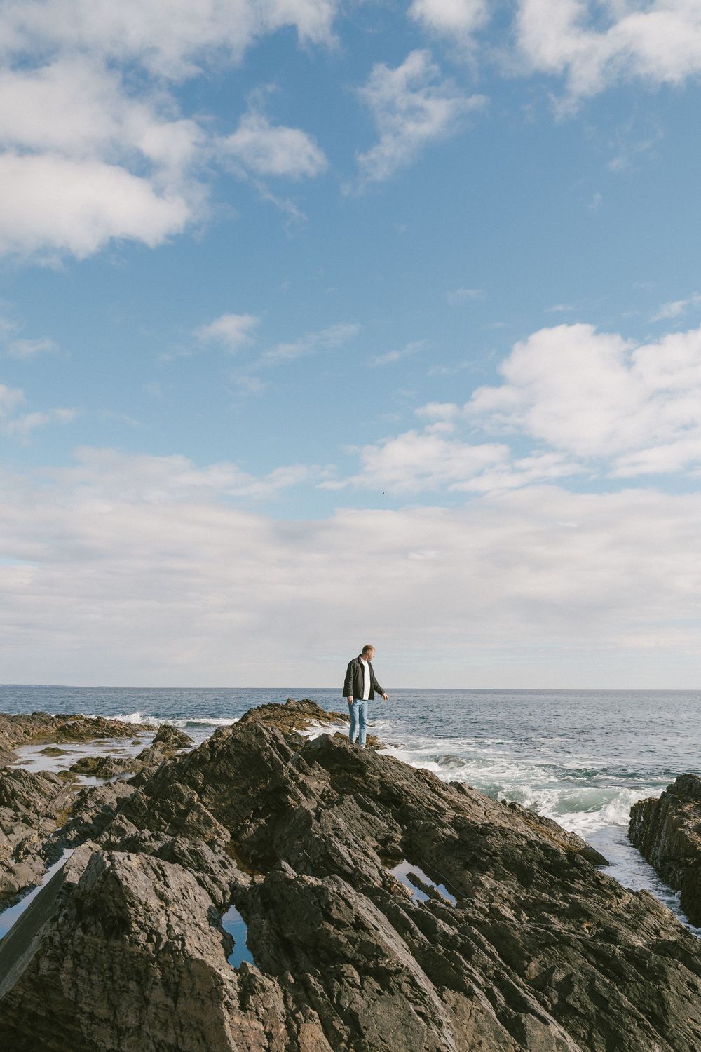 A lone figure stands on rugged coastal rocks overlooking the ocean waves under a bright blue sky with scattered clouds.