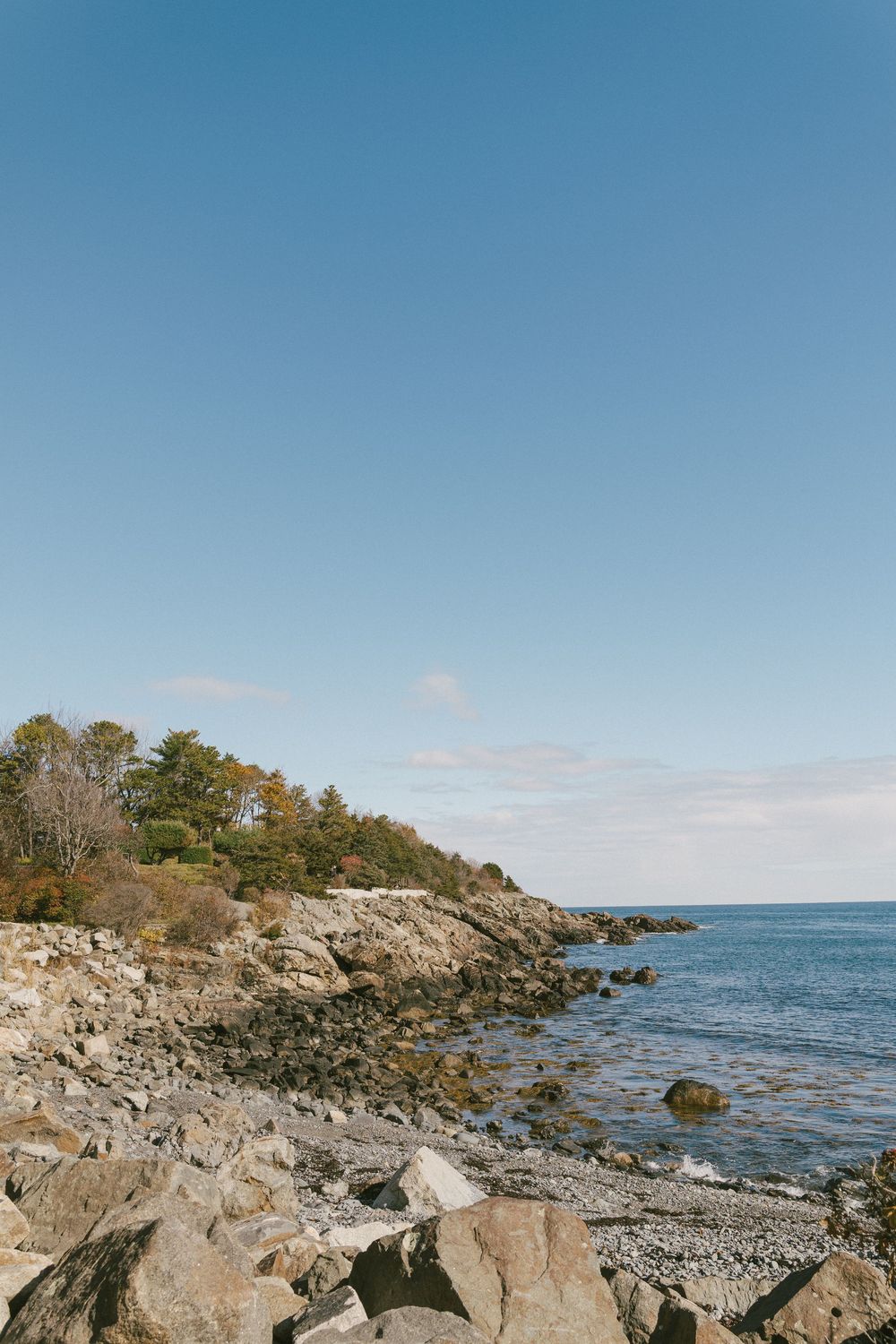 Rocky coastal shoreline with large boulders and calm blue waters under a clear sky at sunset.