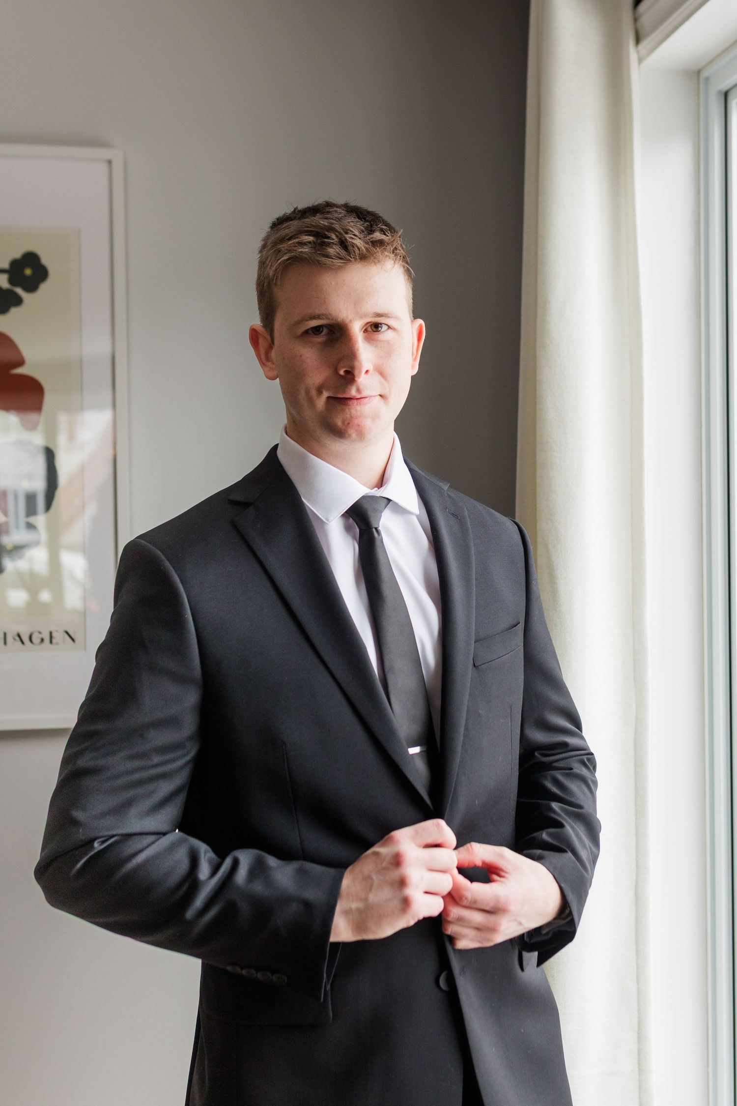 Professional in black suit adjusts his jacket cuff while standing near bright windows in an office setting.