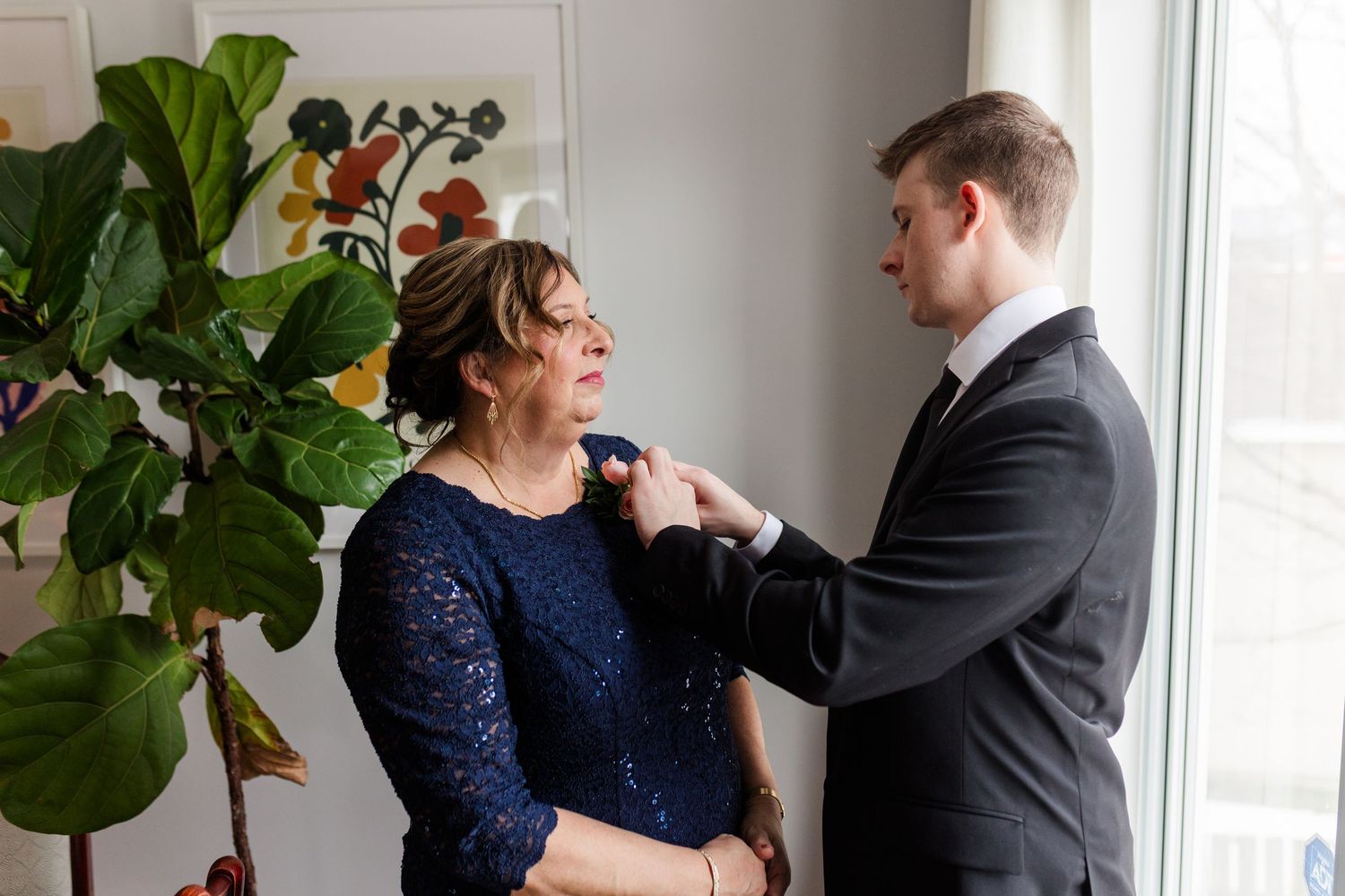 A couple in formal attire shares an intimate moment together by a window with a fiddle leaf fig plant.