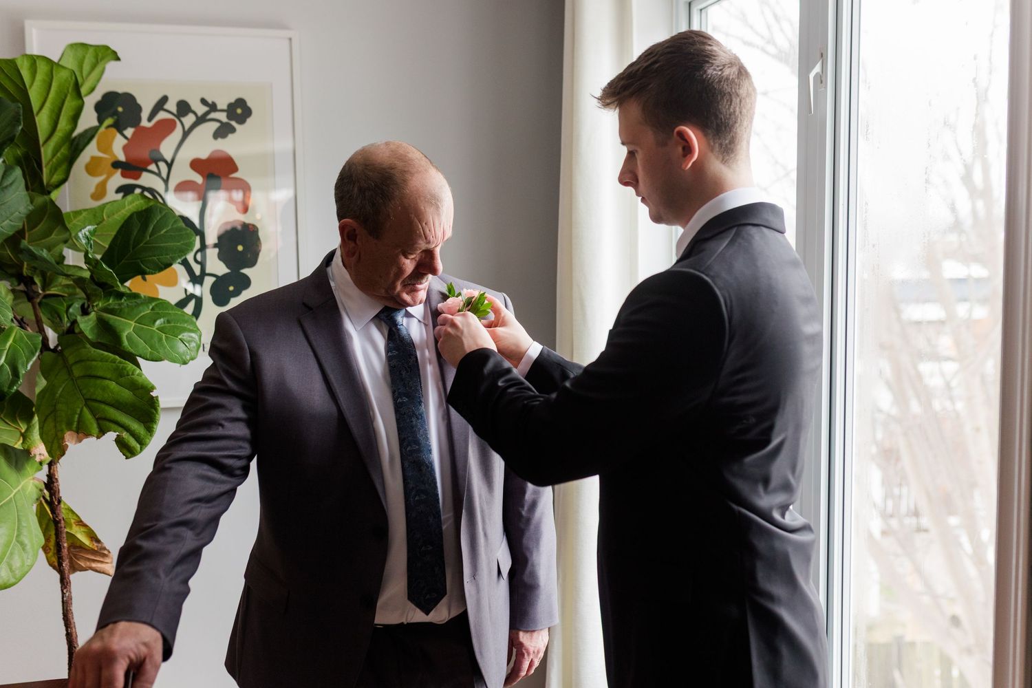 Men in dark suits gather around a table during a business meeting while a fiddle leaf fig plant decorates the room.