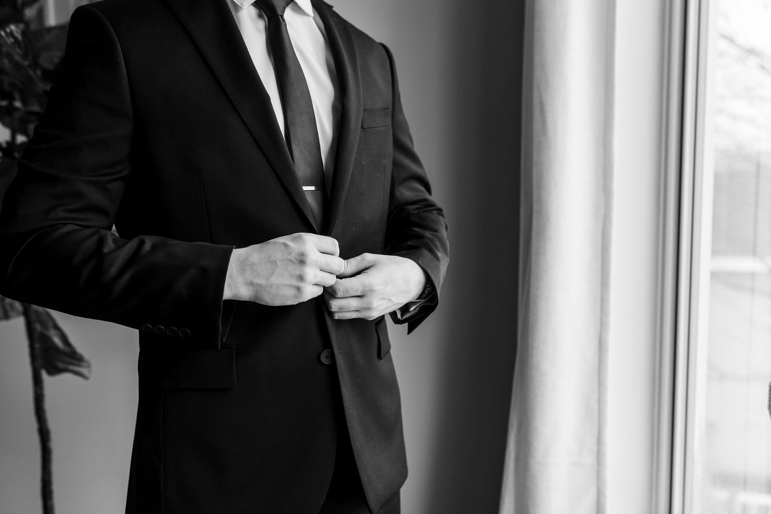 Black and white photo of person in formal suit adjusting tie while standing near window.