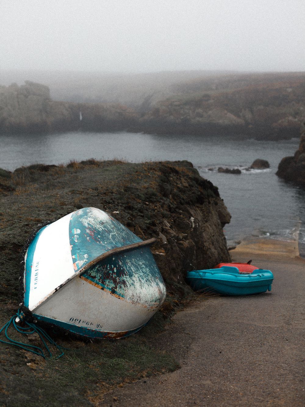 Four overturned boats rest on a misty rocky coastline with dramatic cliffs and ocean waves in the background.