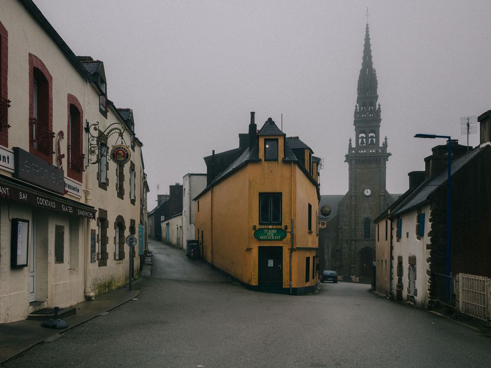 A narrow street in a small European town with a yellow building at the center and a church spire visible in the background.