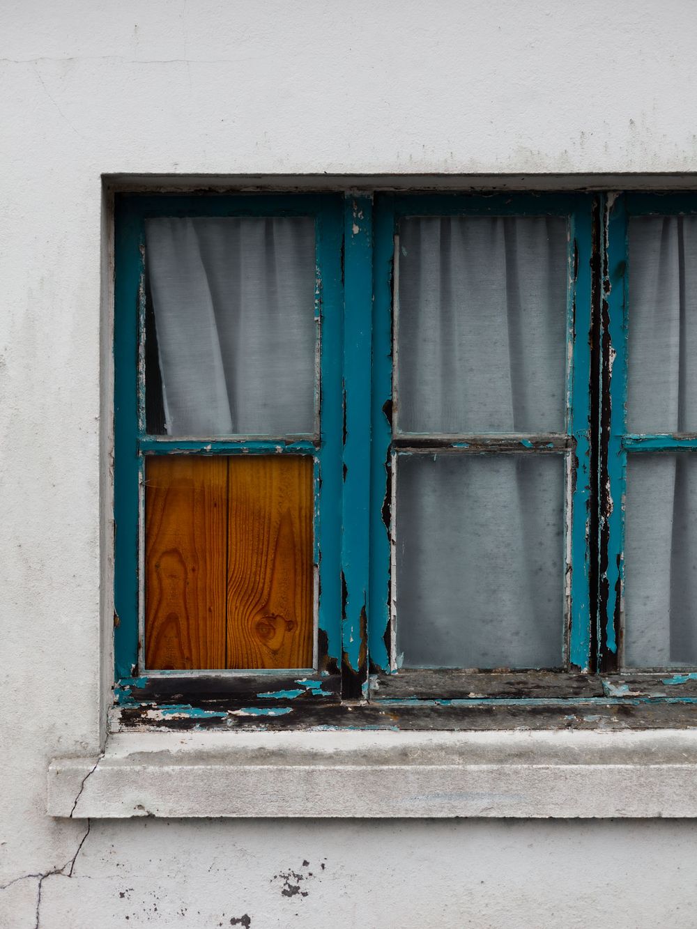 A weathered window frame painted turquoise blue with a wooden panel insert, set against a white wall.