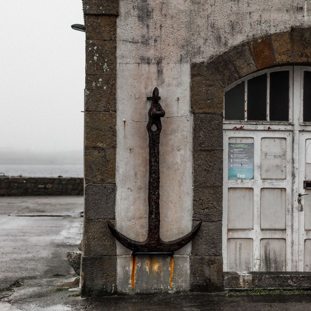 A large rustic anchor mounted on a weathered concrete wall beside a white door in a coastal setting.