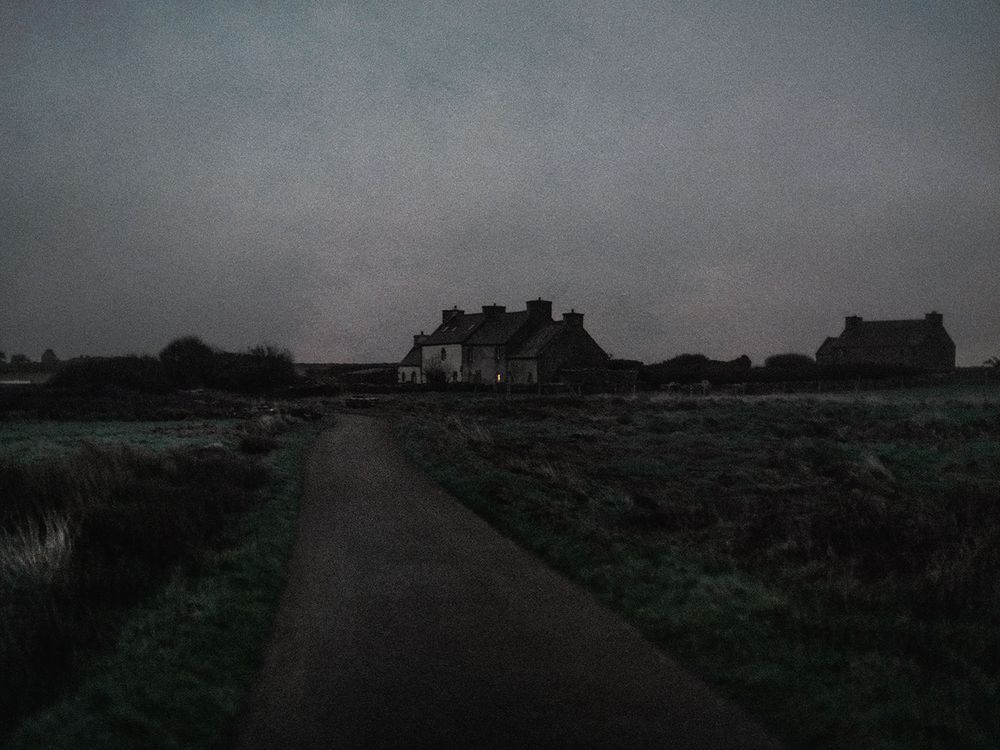 A dark and moody path leads to silhouetted houses on the horizon at dusk in a rural landscape.