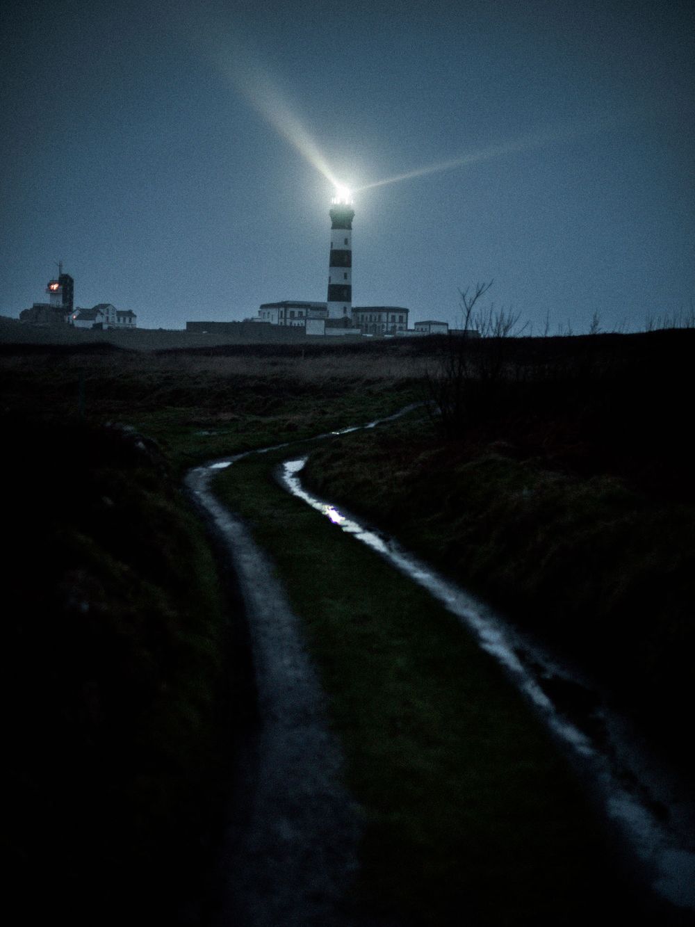 Illuminated lighthouse beacon shines against dark night sky with curved dirt path leading to its base.