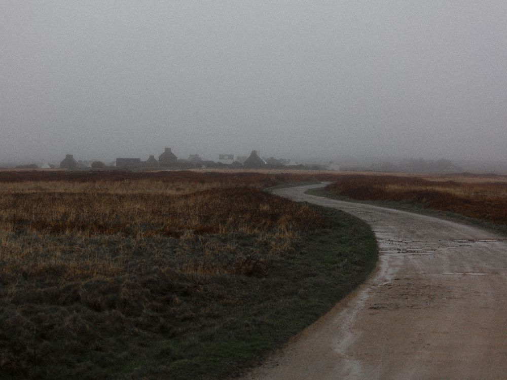 Winding dirt path leads through misty rural landscape with rolling hills and distant buildings on the horizon.