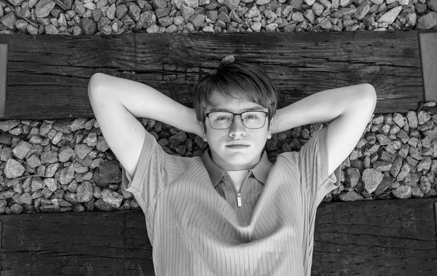 Black and white photo of a young man with glasses lying on a wooden bench with hands behind his head.
