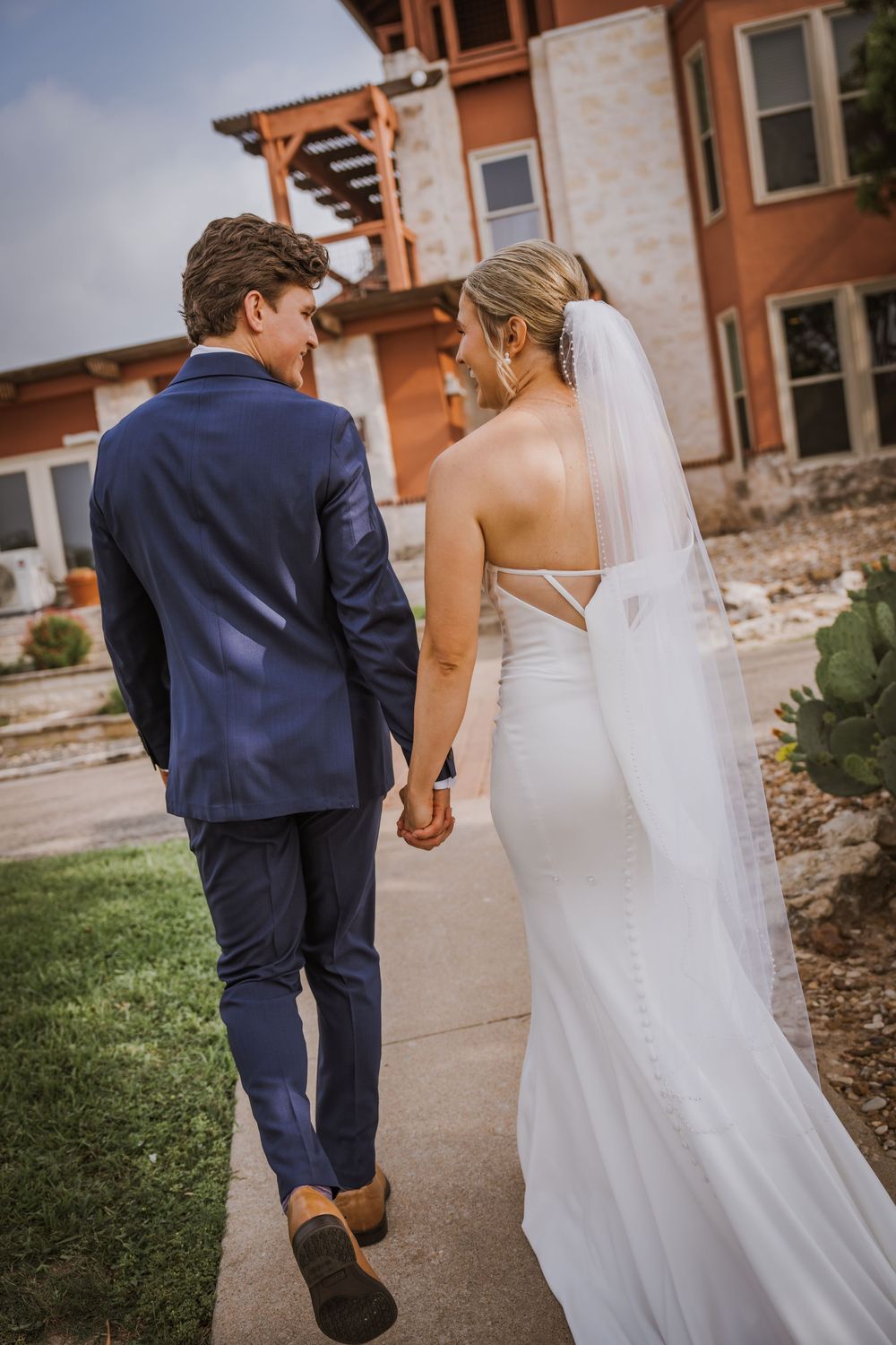 A couple walks hand in hand along a pathway near a terracotta-colored building.