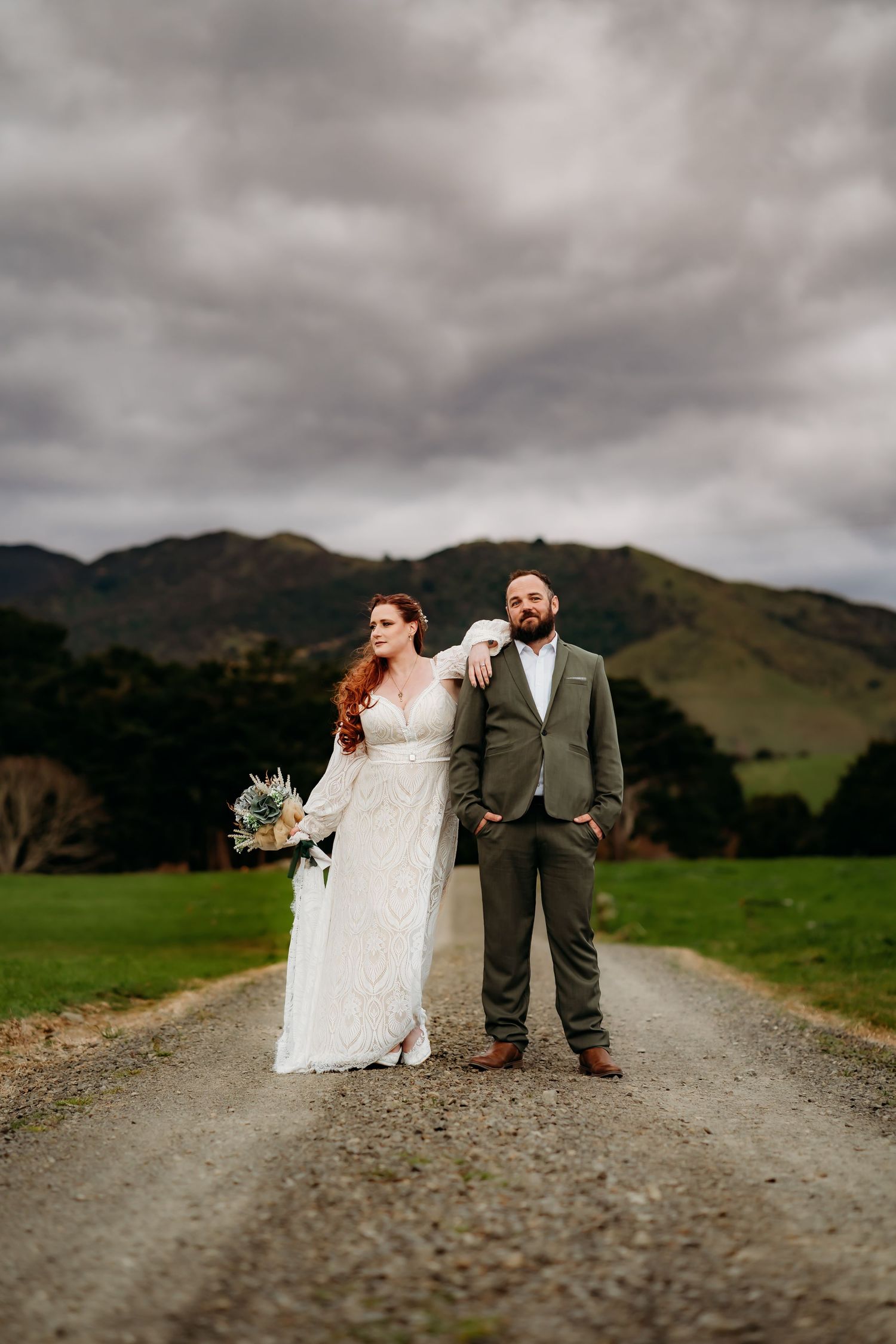 A couple in wedding attire poses on a gravel road with green mountains and moody clouds in the background.