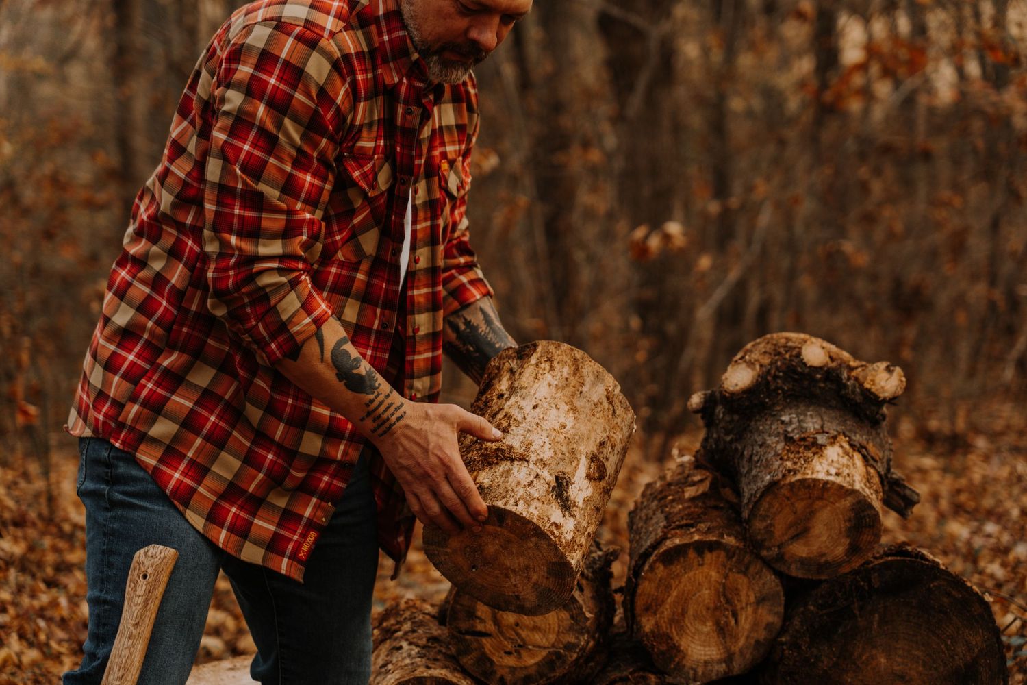 Someone in a red plaid flannel shirt stacks firewood logs in an autumn forest setting.