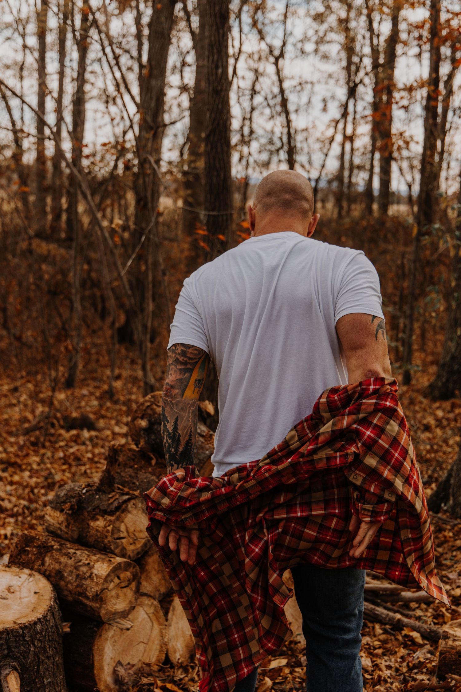 Person in white t-shirt and plaid flannel shirt stands in autumn forest with fallen leaves and bare trees.