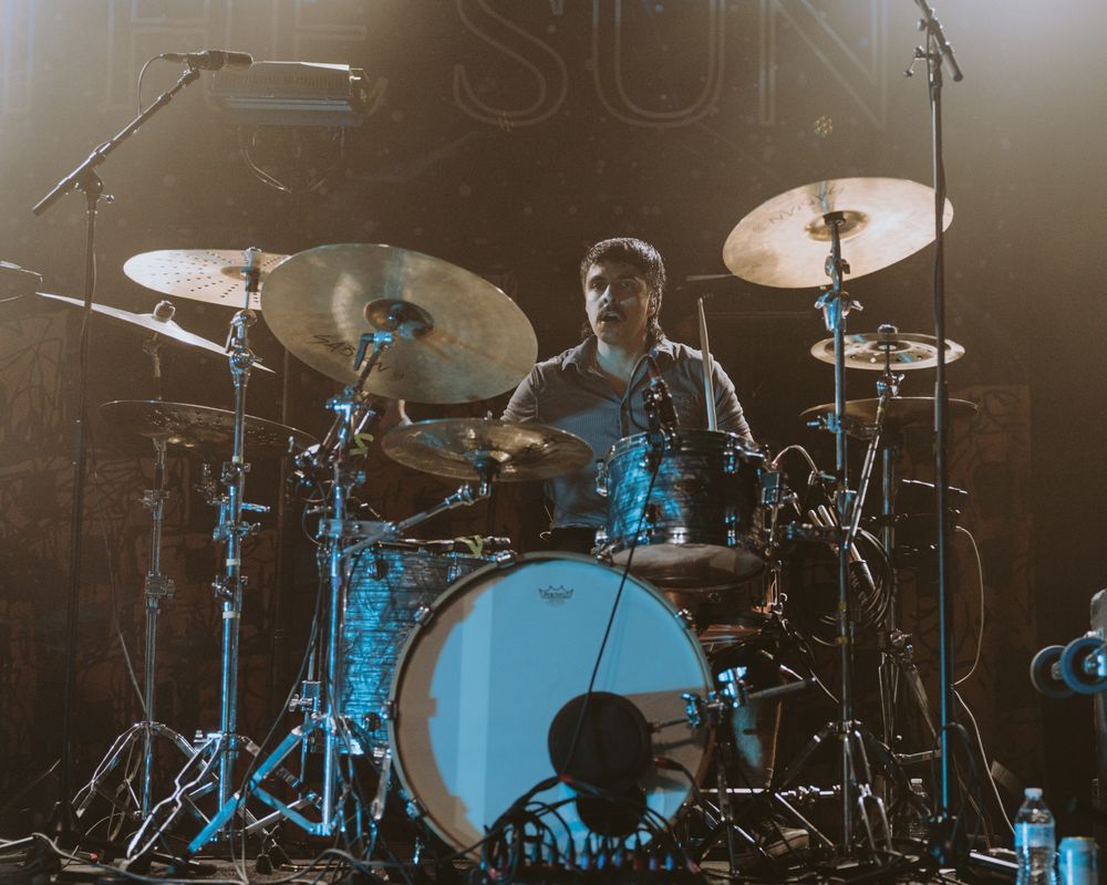 Drummer performs behind illuminated drum kit with blue stage lighting effects.