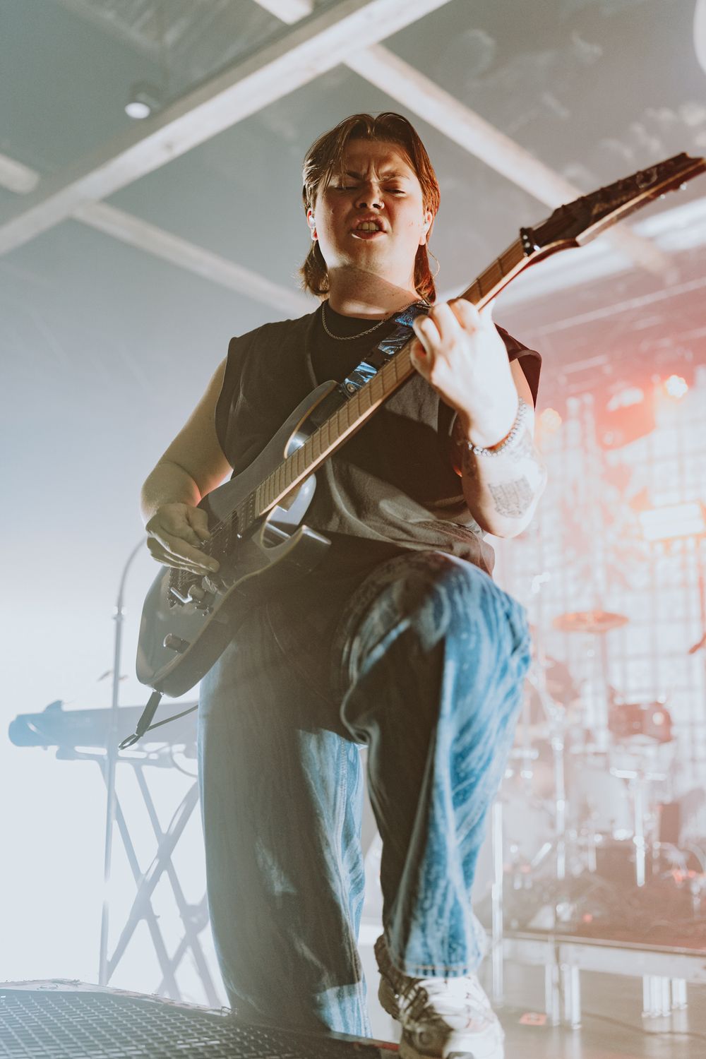 Musician plays guitar on stage wearing casual attire with dramatic backlighting.