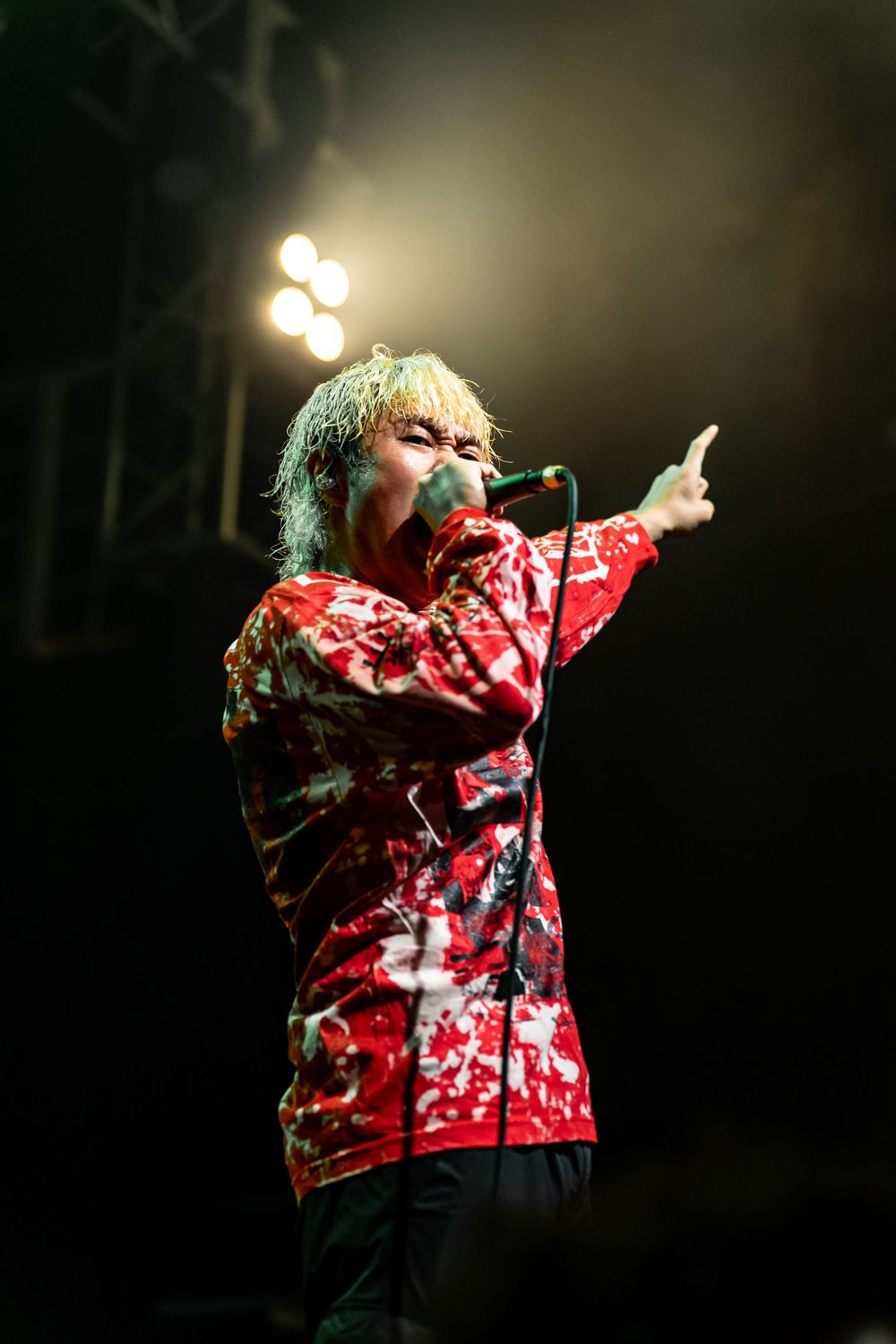 Performer in red patterned jacket singing on dimly lit stage with spotlights.
