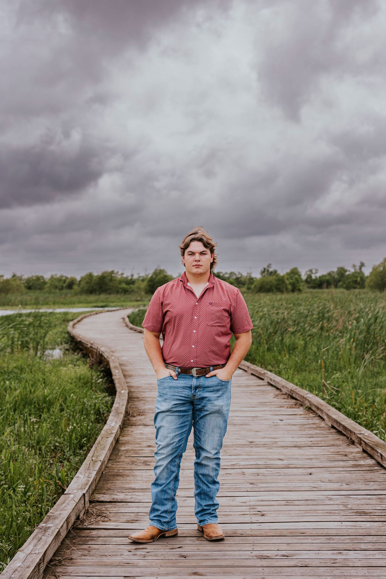 Person in red shirt and jeans stands on wooden boardwalk under stormy sky.
