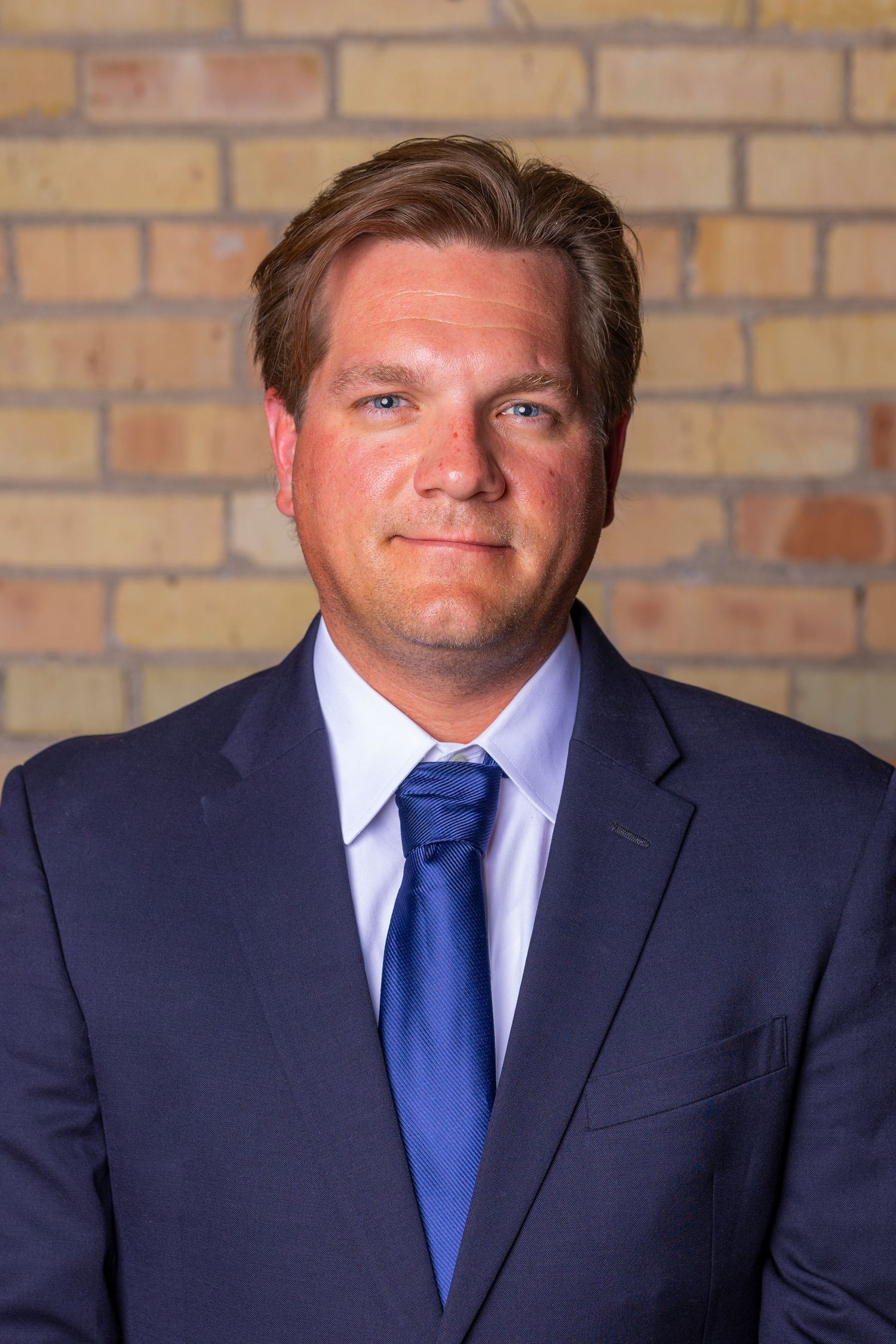 Professional headshot of a person in a navy suit and blue tie standing against a brick wall background.