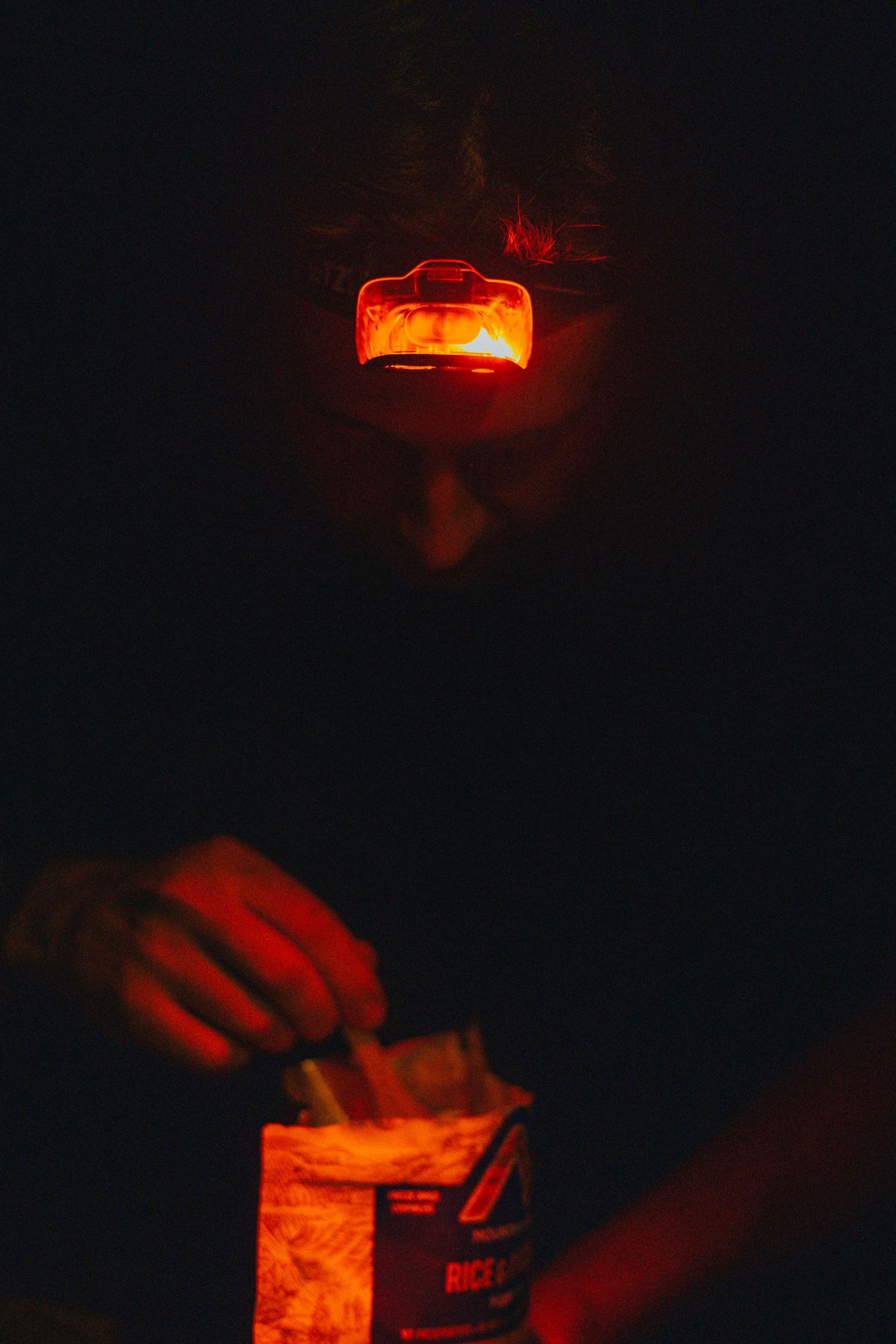 Red headlamp illuminates a dark scene as someone prepares camping supplies at night.