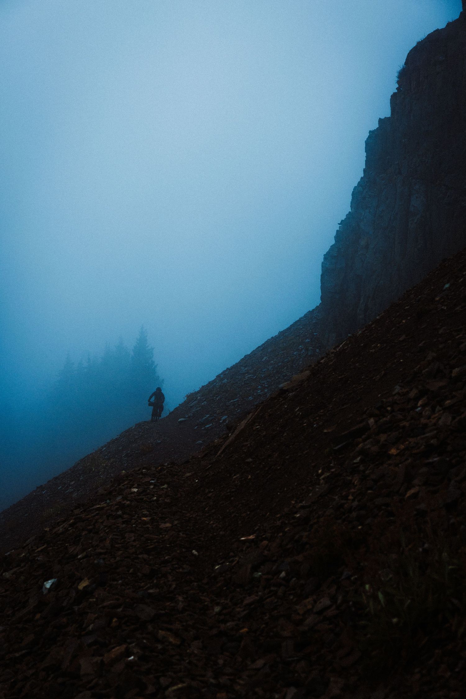 A silhouetted figure stands on a misty mountain slope against a moody blue sky during twilight hours.