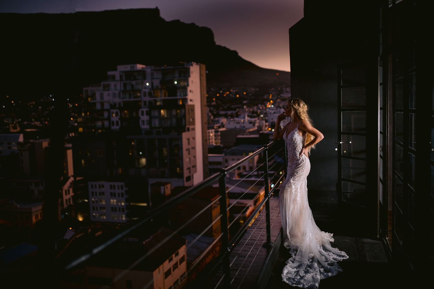 A bride in a white wedding dress stands silhouetted against a cityscape at sunset.