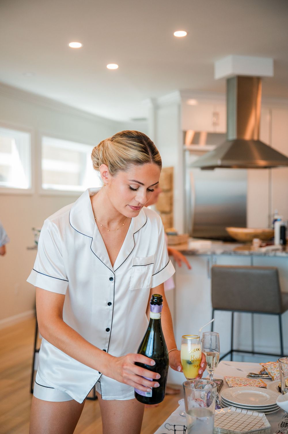 Woman in white pajamas pouring drinks in a modern kitchen during wedding day preparations.