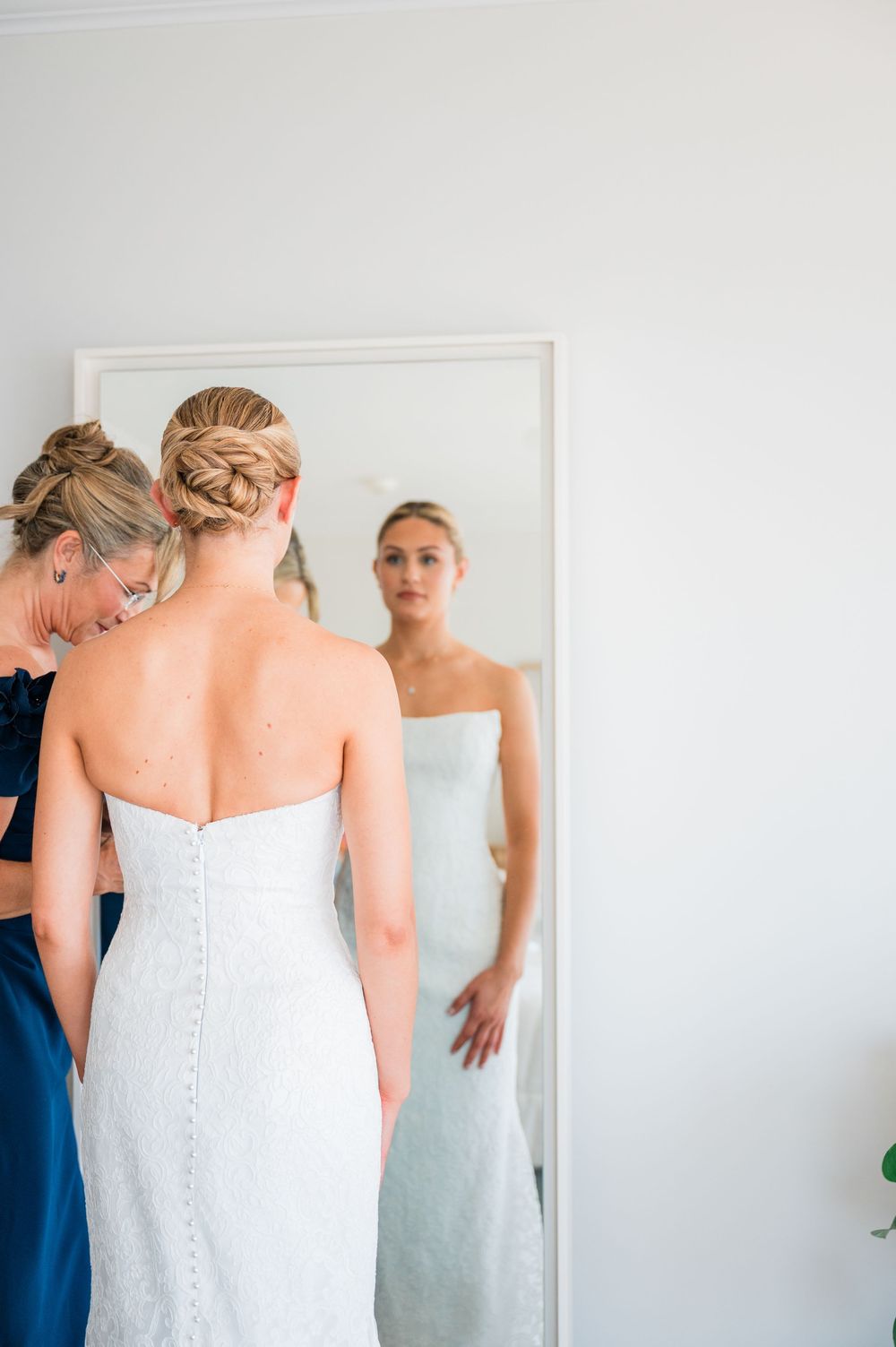 Back view of a white wedding dress being fitted in a bright room with mirrors.