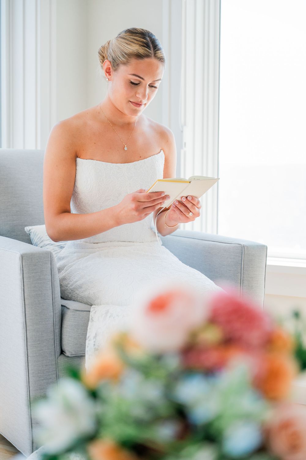 A person in white sits on a grey couch reading a card next to colorful floral arrangements in a bright room.