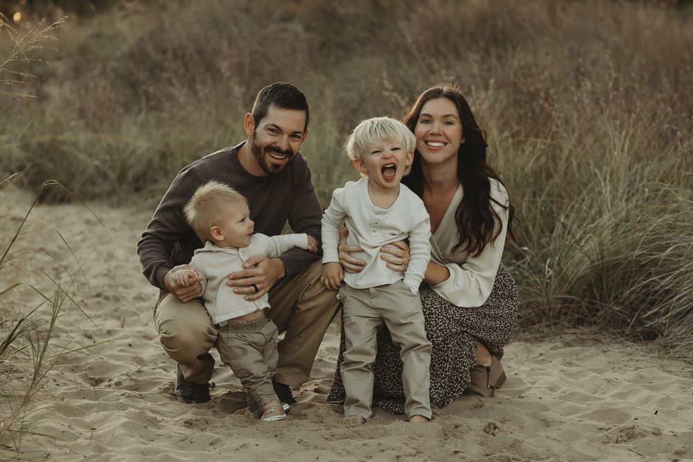 A family sits together on sandy ground surrounded by tall grass during an outdoor photo session at sunset.