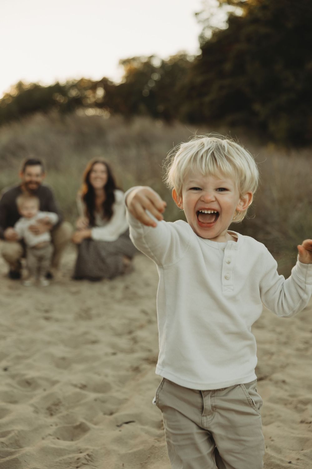 A joyful child wearing a white sweater plays in the sand during a golden hour photoshoot.
