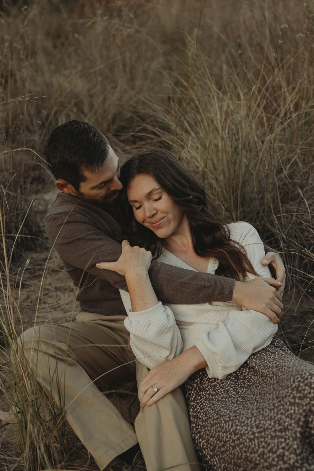 A romantic couple embraces in tall grass during a moody outdoor photoshoot at sunset.