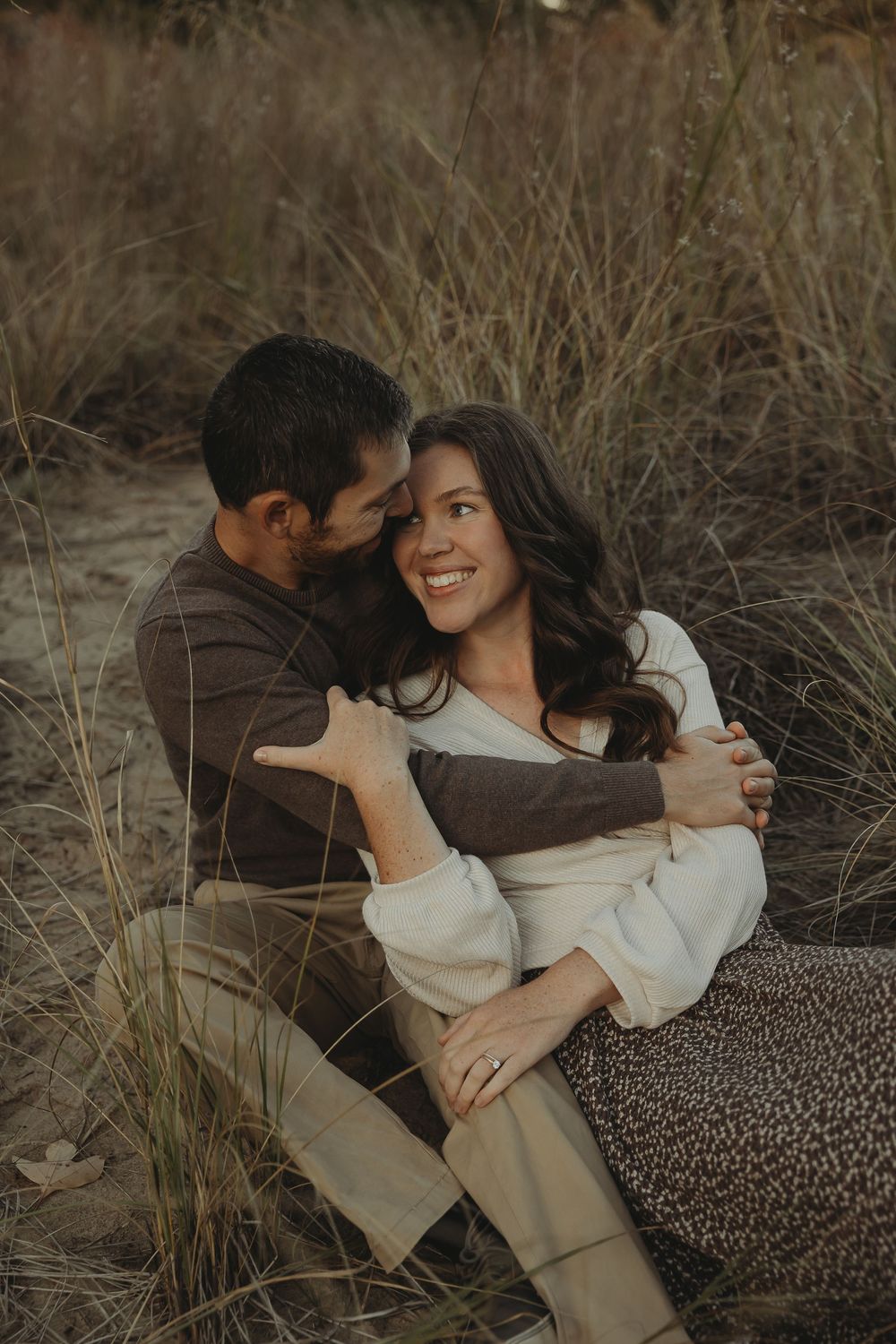 A romantic couple embraces while sitting in tall grass during a moody outdoor photo session.