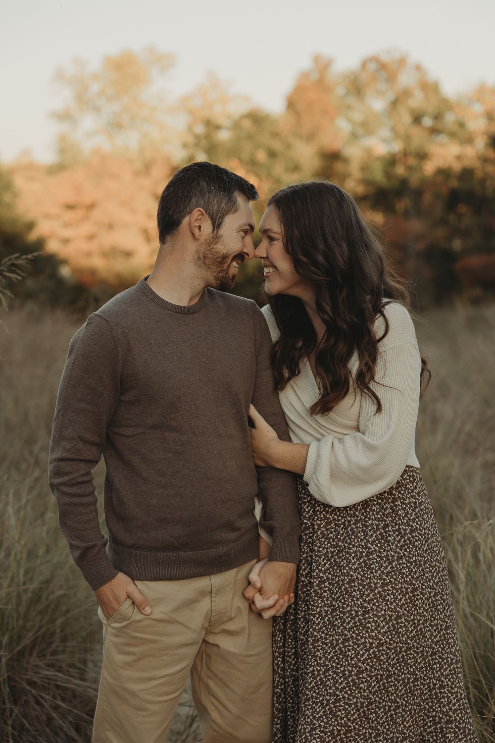A romantic couple embraces in a natural field setting during sunset wearing brown and cream colored outfits.