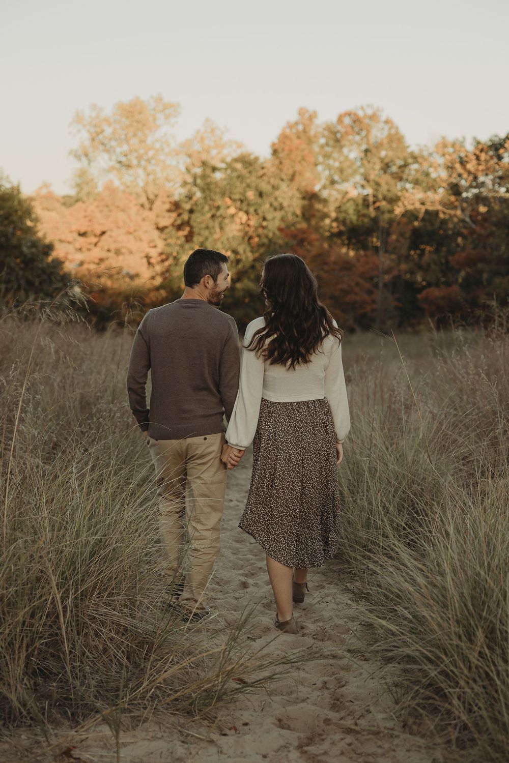A couple walks together through tall grass at sunset in a romantic outdoor setting.
