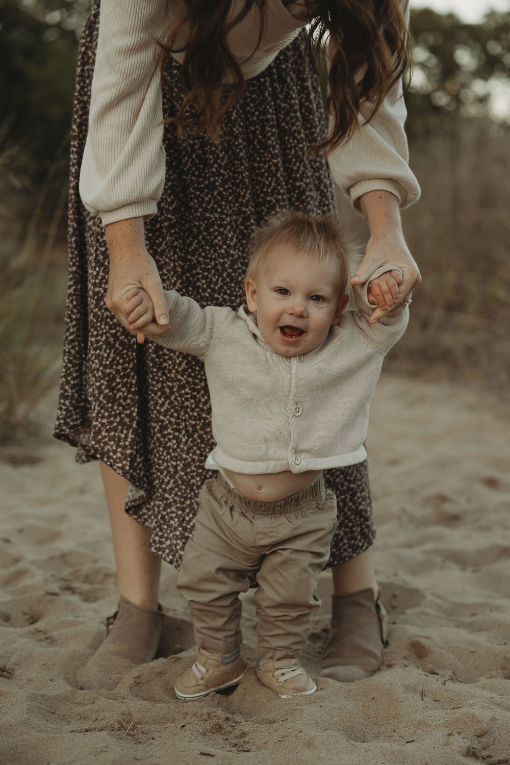 A baby in a cream sweater smiles while learning to walk on sand, supported by someone in a floral dress.