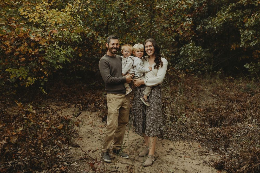 A group of people in coordinated fall outfits pose together in a wooded setting during autumn.