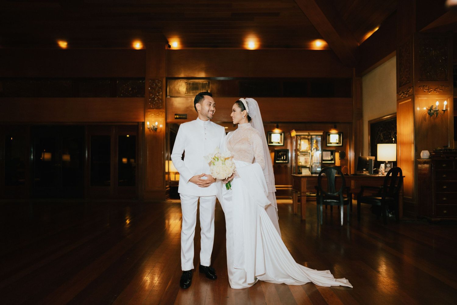 Elegant bride and groom in white attire share a romantic moment in a warmly lit wooden ballroom.