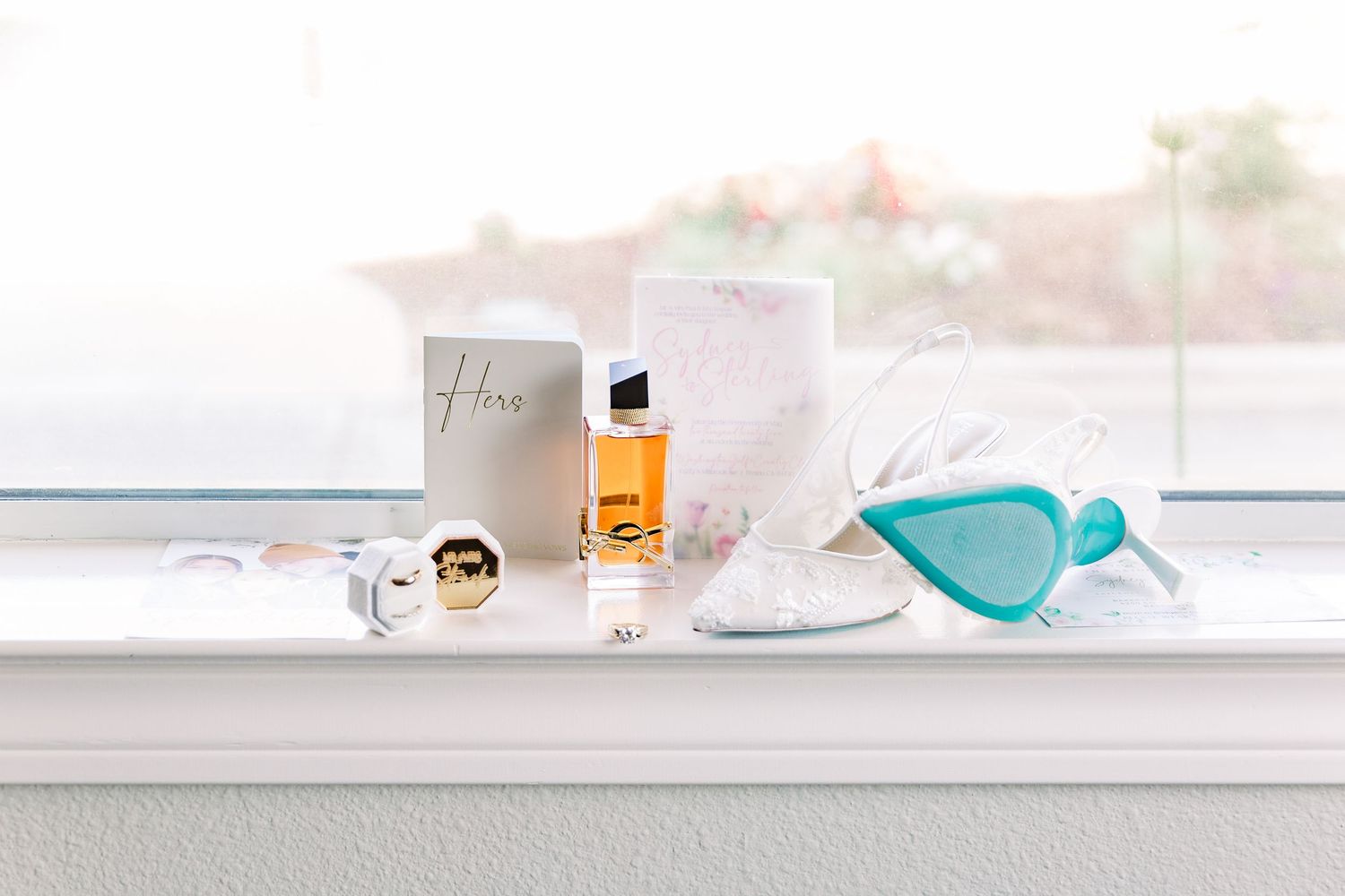 Wedding day details including perfume, sunglasses and invitation displayed on white ledge.