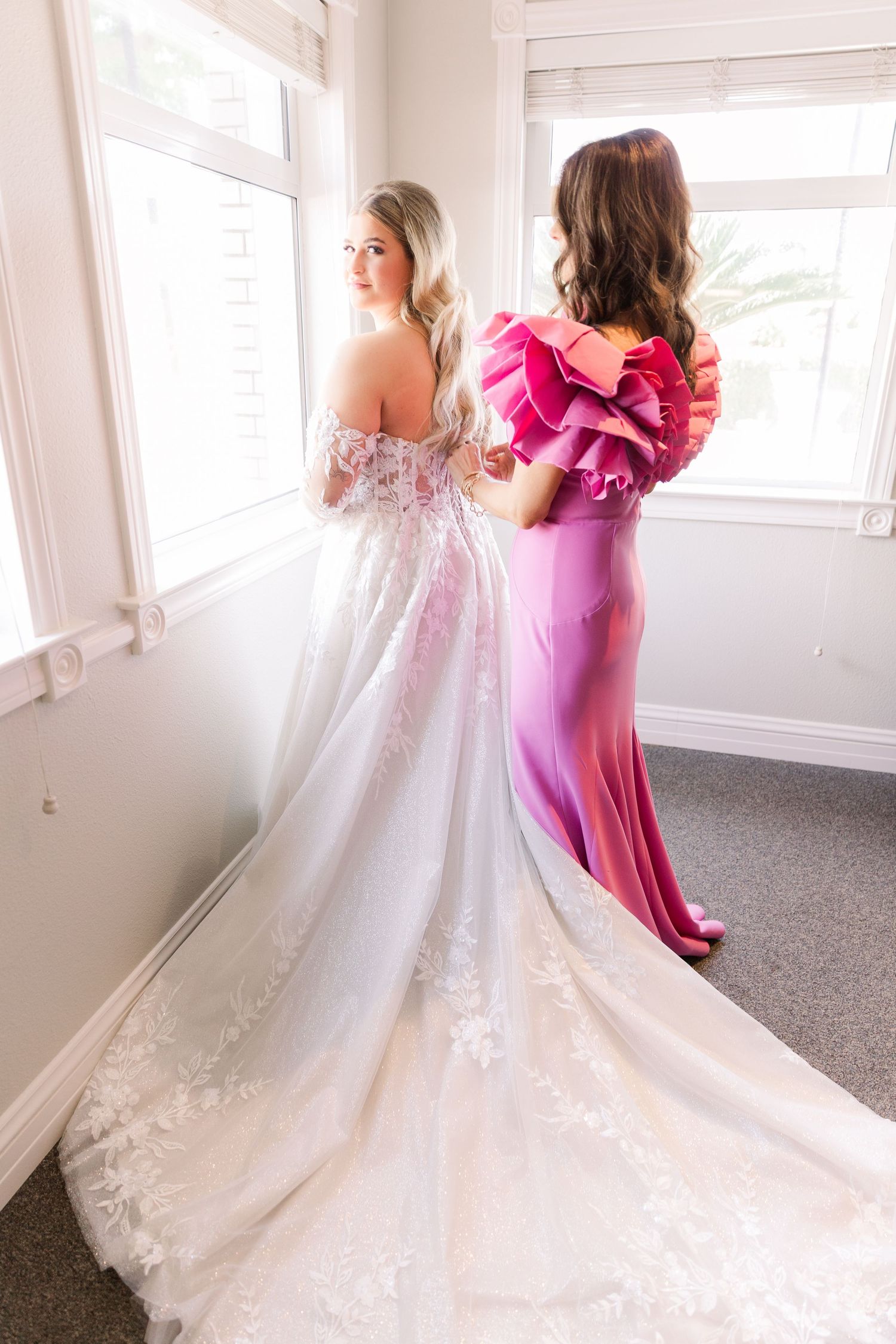 Two women in formal dresses stand by a window, one in white and one in bright pink.