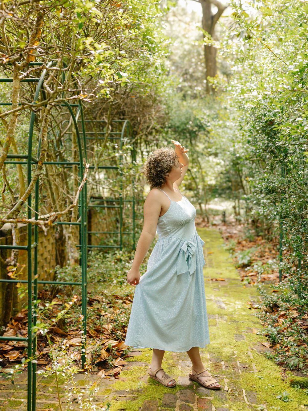 Whimsical poses in a light blue dress along a garden pathway.