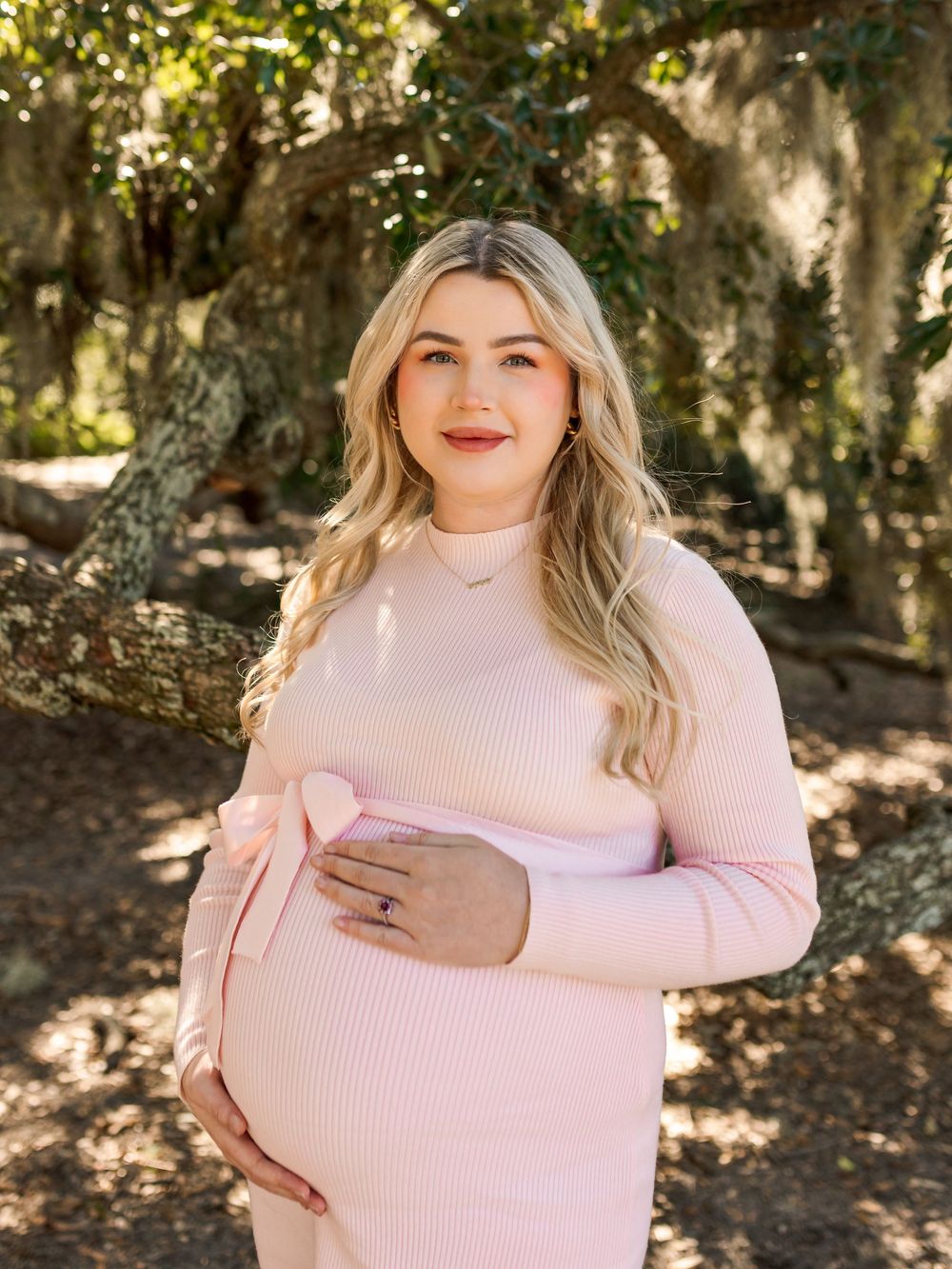 Sunlit maternity portrait in a natural outdoor setting with moss-draped trees.